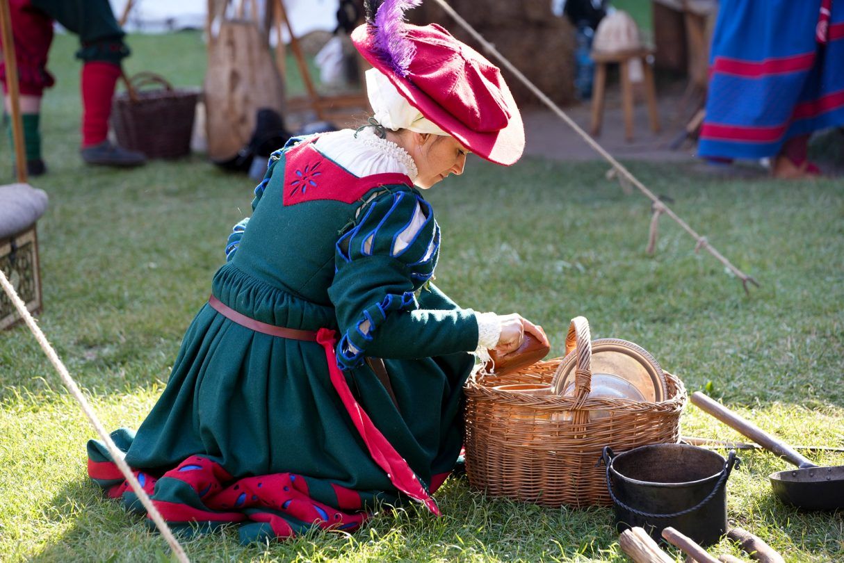 kobieta w kolorowym średniowiecznym stroju siedzzi na trawie i spogląda do koszyka
